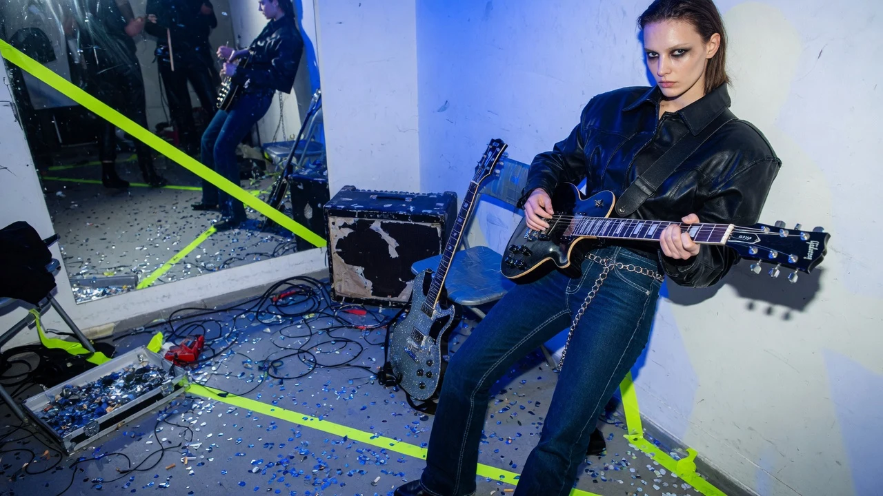 High-fashion editorial still inside an intimate rehearsal space under direct flash with guitars, confetti, and electric blue light.
