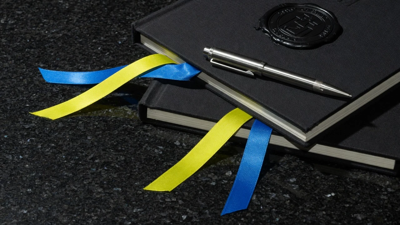 A closed leather portfolio folder and a blue ceramic vessel on a walnut office desk in window light.