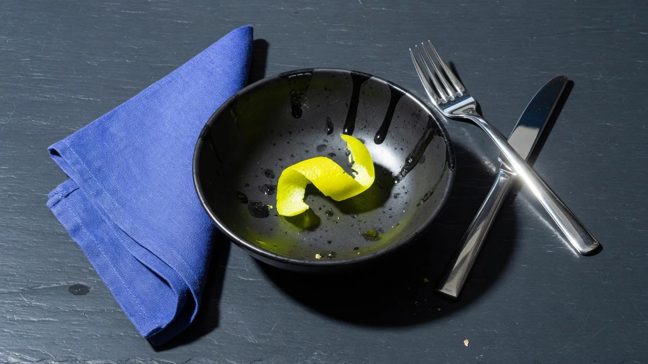 A stoneware plate with a linen napkin and rosemary, and a blue ceramic vessel on a walnut office desk in window light.