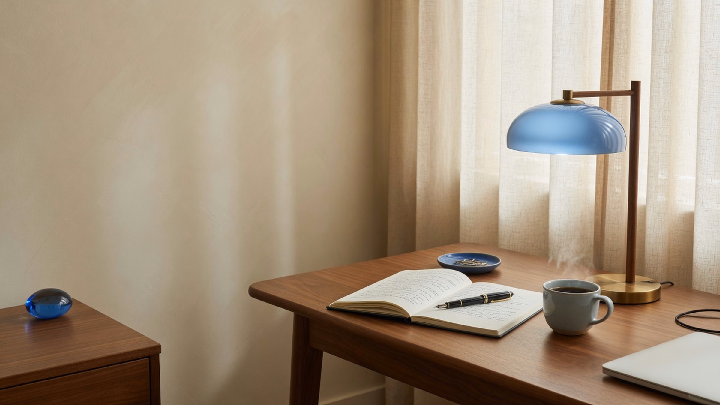 Walnut writing desk with open notebook, fountain pen, coffee, closed laptop edge; #1B6BB5 glass lamp shade and small blue dish; cream plaster and linen curtain; left third calm