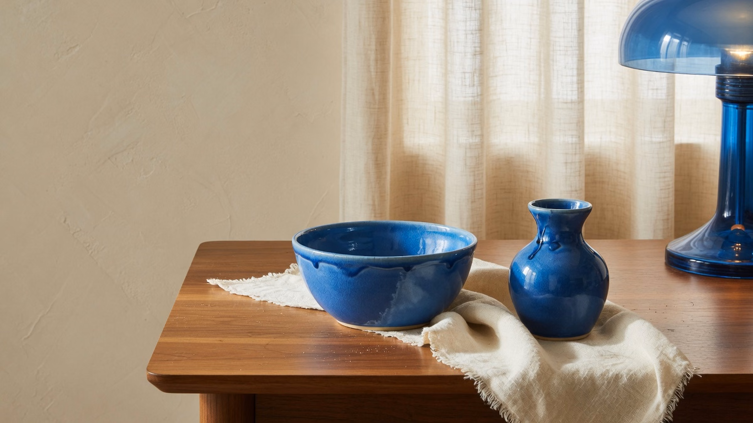 Glossy #1B6BB5 ceramic bowl and bud vase on rumpled linen over walnut desk; cream plaster and linen curtain; blue glass lamp edge; no people