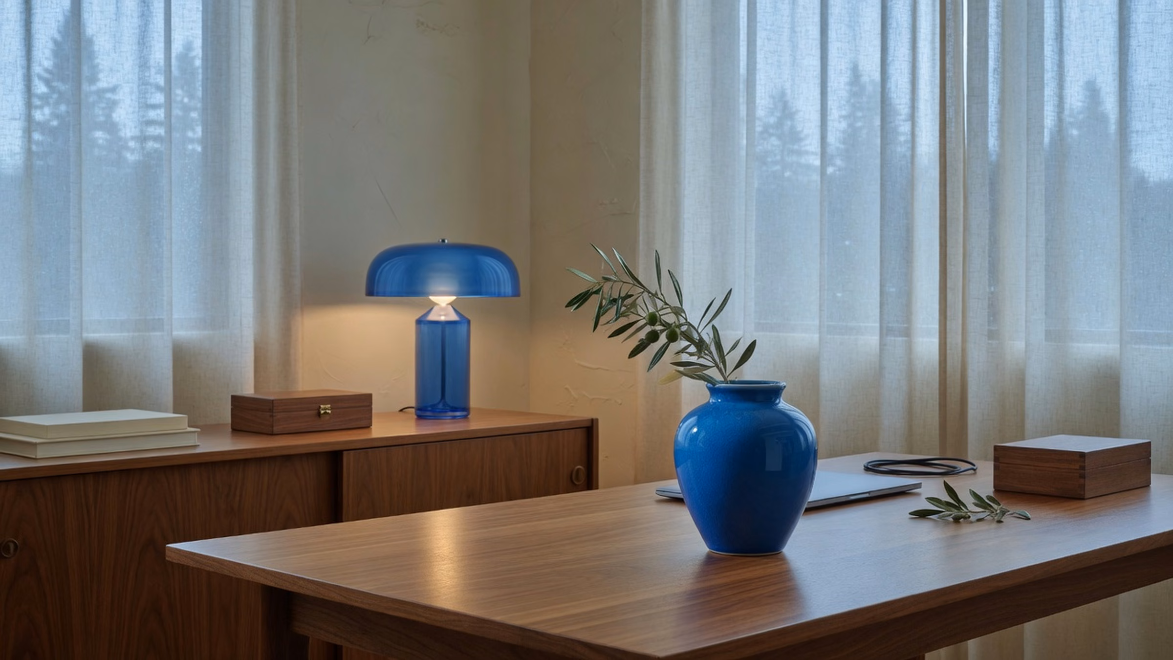 Close-to-mid warm walnut desk and cream plaster with sheer linen; glossy #1B6BB5 ceramic vessel with olive sprig and blue glass lamp; no people, no readable screens